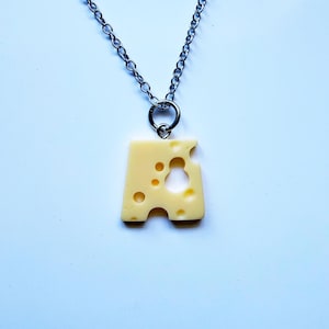 May include: A silver-toned metal chain necklace featuring a pendant shaped like a wedge of pale yellow Swiss cheese. The cheese pendant has several circular holes of varying sizes cut out of its surface and is attached to the chain by a small ring.