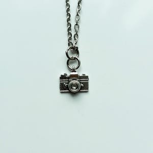 May include: A silver-colored camera pendant necklace. The pendant is a miniature replica of a vintage camera, with detailed features. The necklace chain is also silver. The words "Pissy Prints" are written in cursive at the bottom.