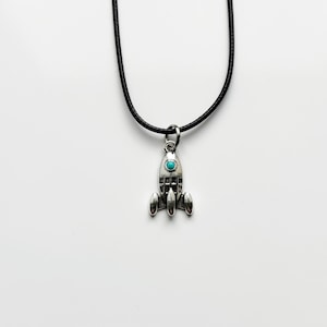 May include: A silver-colored rocket pendant necklace with a turquoise stone set in the center. The rocket has three fins and is suspended from a black cord. The necklace is displayed against a white background.