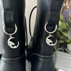 May include: A pair of black leather boots with silver moon and wolf charms attached to the laces. The boots have a chunky platform sole and a lace-up front.