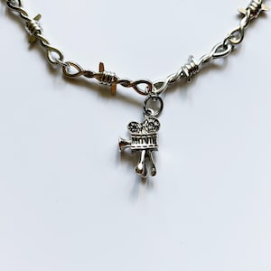 May include: A silver-toned necklace featuring a chain designed to resemble barbed wire. A small, detailed silver charm shaped like a vintage movie camera hangs from the center of the chain, with the word MOVIE clearly embossed on its side.