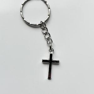 May include: A silver-colored keychain with a small cross pendant. The cross is attached to the keychain ring by a short chain. The background is plain white.