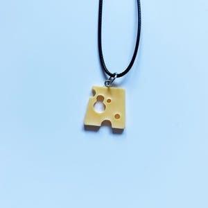 May include: A necklace featuring a pale yellow, triangular cheese-shaped pendant with several circular holes, suspended from a thin black cord by a small silver-toned metal jump ring. The pendant mimics the appearance of Swiss-style cheese.