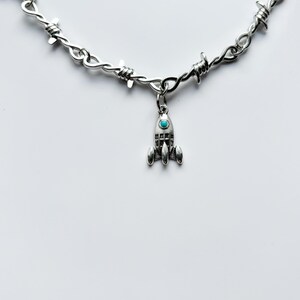 May include: Silver-toned necklace featuring a barbed wire chain design. A small rocket pendant with a turquoise-colored stone hangs from the chain. The necklace is displayed against a white background.