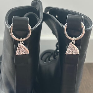 May include: Black leather boots with silver-toned pizza slice charms hanging from the top loops. The charms have detailed patterns. The boots are laced up and have a classic combat boot style.
