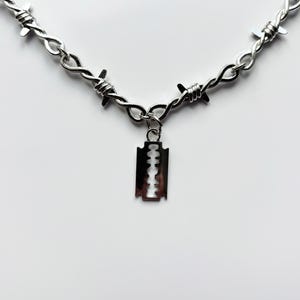 Könnte beinhalten: Eine silberfarbene Halskette mit einem Stacheldraht-Design. Ein kleiner, silberner Rasierklingen-Anhänger hängt von der Mitte der Halskette. Die Halskette wird vor einem weißen Hintergrund gezeigt.