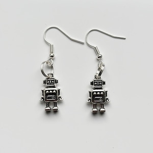 May include: A pair of silver-colored robot earrings. Each earring features a small, detailed robot charm hanging from a silver fish hook. The robots have rectangular bodies, small arms, and legs, with detailed head and chest features.