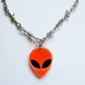 May include: A necklace featuring a silver-toned chain designed to resemble barbed wire. The pendant is a bright neon orange alien head shape with two large, smooth, black oval-shaped eyes set into the center of the orange material.