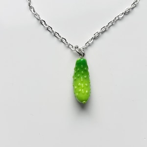 May include: A silver chain necklace featuring a green pickle pendant. The pickle pendant is a vibrant green with a textured surface, suspended from a small silver loop. The chain is a simple link design.