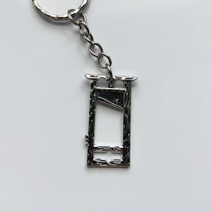 Peut inclure: Un porte-clés argenté représentant une guillotine miniature. Le porte-clés comprend un anneau et une chaîne pour l'attacher à des clés ou à d'autres objets. La guillotine a un aspect détaillé et texturé.