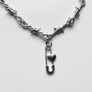 Può includere: Collana color argento con un design a filo spinato. Un ciondolo a spilla da balia con un ciondolo a cuore pende al centro. La collana è mostrata su uno sfondo bianco, evidenziando la finitura metallica.