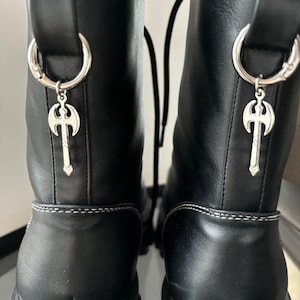 Puede incluir: Botas de cuero negro con abalorios de hacha plateada unidos a los cordones. Las botas tienen una suela de plataforma gruesa y un cierre de cordones.