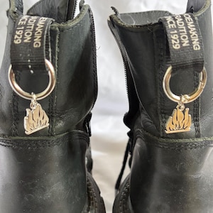 May include: A pair of black leather boots featuring a side zipper and a black pull tab at the heel with the text "SHOEMAKING TRADITION SINCE 1929". A silver-toned metal ring is attached to each pull tab, holding a silver flame-shaped charm.