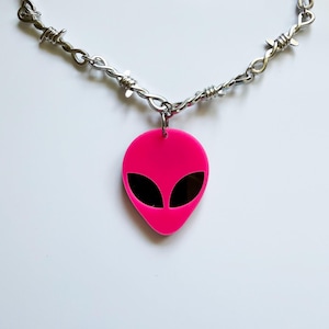 May include: A silver-toned necklace featuring a chain designed to resemble barbed wire. The pendant is a bright neon pink alien head shape with two large, smooth, black oval-shaped eyes centered on the face. The pendant hangs from a metal jump ring.