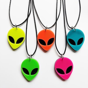 May include: Five necklaces featuring alien head pendants in neon yellow, orange, teal, lime green, and hot pink. Each pendant has black cutout eyes and is suspended from a thin black cord. The alien shapes are smooth and brightly colored.