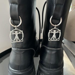 May include: A pair of black leather boots with silver charms attached to the laces. Each charm features a silver outline of a human figure with outstretched arms.