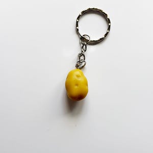 May include: A yellow potato-shaped keychain with a silver-colored ring and chain. The potato charm has a textured surface and a small, dark spot. The keychain is designed to be attached to keys or bags.