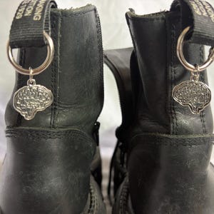 May include: A pair of black leather boots featuring silver-toned brain-shaped charms attached to the heel pull tabs with metal rings. The pull tabs have black fabric straps with white text that reads MAKING TRADITION SINCE 1929.