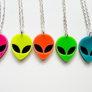 May include: Five alien head necklaces in vibrant colors: pink, neon yellow, orange, green, and teal. Each pendant has a black eye design and hangs from a silver chain. The pendants are displayed against a white background.
