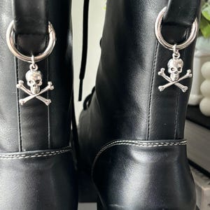 Puede incluir: Botas de cuero negro con costuras blancas y abalorios de calavera y huesos cruzados unidos a los cordones.