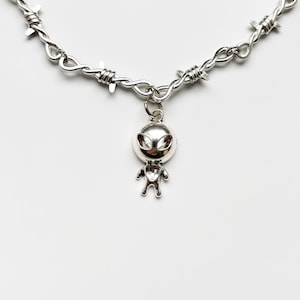 May include: Silver-toned necklace featuring a barbed wire design. A small, detailed alien pendant hangs from the center. The alien has a rounded head and small arms and legs. The necklace is set against a white background.