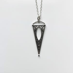 May include: A silver-toned pendant necklace with a triangular, openwork design. The pendant has an elongated oval center and intricate patterns. The chain is a delicate silver color, and the necklace is displayed against a white background.