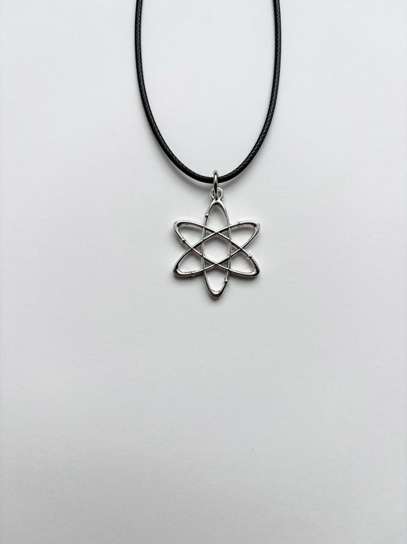 以下が含まれることがあります： 銀色の原子ペンダントネックレス。ペンダントは、六芒星の形をした原子モデルです。ネックレスは黒いコードで、白い背景にセットされています。