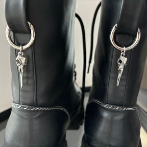 Puede incluir: Un par de botas de cuero negro con costuras blancas y dijes de calavera plateados unidos a los cordones.