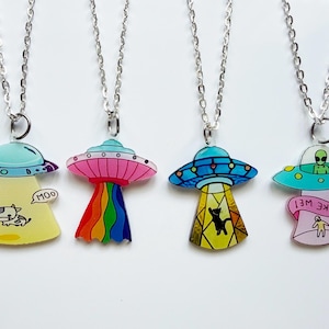 May include: Four silver chain necklaces with colorful UFO pendants. Each pendant features a unique design: a cow with "MOO", a rainbow beam, a cat, and an alien with "TAKE ME!" text. The pendants are in shades of yellow, pink, blue, and green.