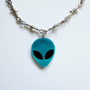 May include: A silver-toned metal chain necklace featuring a repeating barbed wire design. A single, flat, bright turquoise alien head pendant with two large, smooth, black oval-shaped eyes is attached to the center of the chain by a small metal ring.
