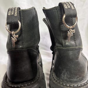 Könnte beinhalten: Ein Paar schwarze Leder-Plateaustiefel mit dicken Sohlen, seitlichen Reißverschlüssen und schwarzen Zuglaschen mit dem Text "MAKING DITION CE 1929". Jeder Stiefel hat einen silberfarbenen Metallring an der Zuglasche mit einem kleinen Revolver-Anhänger.