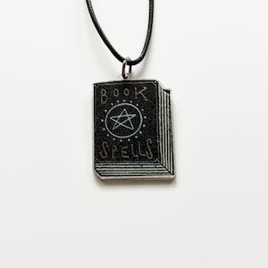 Peut inclure: Un collier pendentif noir avec un charme en forme de livre. Le charme porte les mots "BOOK SPELLS" et un pentagramme. Le pendentif est suspendu à un cordon noir sur fond blanc.