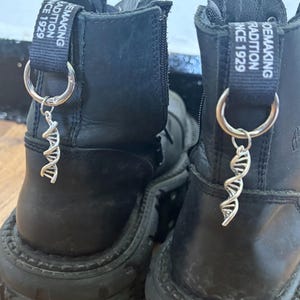 Könnte beinhalten: Schwarze Lederstiefel mit silbernen DNA-Helix-Anhängern. Die Stiefel haben dicke Sohlen und die Zuglaschen tragen die Aufschrift "Shoemaking Tradition Since 1929".