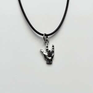 Può includere: Collana con pendente color argento raffigurante un gesto della mano con l'indice e il mignolo estesi. Il pendente è sospeso a un sottile cordino nero. Lo sfondo è una superficie bianca.