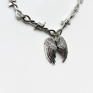 May include: A silver-coloured necklace with a barbed wire design. The necklace has a pair of detailed wing charms hanging from the centre. The wings are intricately designed with feather details. The background is plain white.