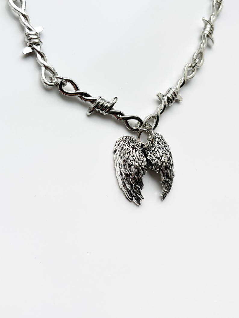May include: A silver-coloured necklace with a barbed wire design. The necklace has a pair of detailed wing charms hanging from the centre. The wings are intricately designed with feather details. The background is plain white.