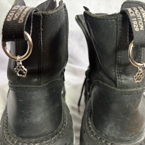 Könnte beinhalten: Ein Paar schwarze Lederstiefel mit seitlichem Reißverschluss und einer schwarzen Zugschlaufe an der Ferse mit dem weißen Text "SHOEMAKING TRADITION SINCE 1929". An jeder Schlaufe hängt ein silberner Metallring mit einem Pfotenanhänger.