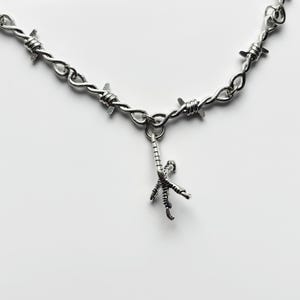May include: A silver-toned necklace featuring a barbed wire design. A small, detailed charm of a bird's foot hangs from the center. The necklace is set against a plain white background.