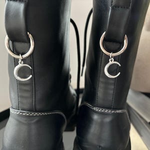 Può includere: Un paio di stivali in pelle nera con cuciture bianche e ciondoli a forma di mezzaluna d'argento attaccati ai lacci.
