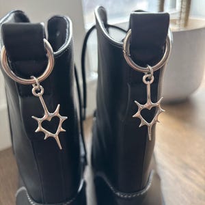 May include: A pair of black leather boots with silver-toned heart-shaped charms hanging from the top. The boots have a chunky sole and are laced up. The charms are attached to silver hoops.