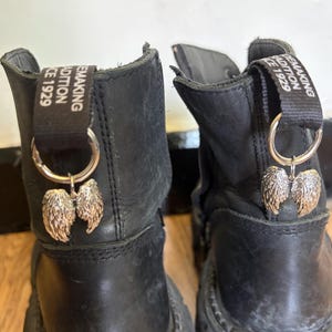 Può includere: Un paio di stivali in pelle nera con una linguetta nera sul retro di ogni stivale con il testo bianco SHOEMAKING TRADITION SINCE 1929. Un anello in metallo argentato è fissato a ogni linguetta, con un piccolo ciondolo a forma di ala.