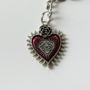 May include: Silver keyring with a red heart-shaped charm featuring a stylised eye and a rose detail.