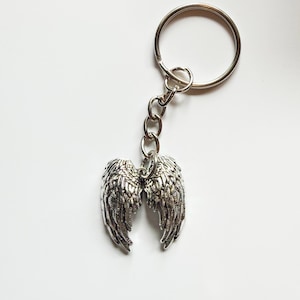 May include: A silver-toned metal keychain featuring a pair of detailed angel wings as the pendant. The wings are attached to a standard circular split ring by a short metal chain, showcasing intricate feather textures and a polished metallic finish.