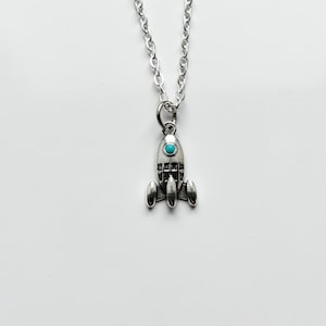 May include: Silver-toned rocket ship pendant necklace with a turquoise-colored stone set in the center of the rocket. The necklace has a silver chain and a simple clasp. The rocket has three fins and a detailed design.