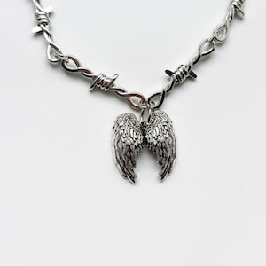 May include: Silver-coloured necklace with a barbed wire chain and a pendant of detailed angel wings. The wings are intricately designed, adding a gothic or edgy aesthetic. The necklace is displayed against a white background.