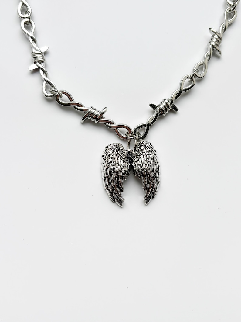 May include: Silver-coloured necklace with a barbed wire chain and a pendant of detailed angel wings. The wings are intricately designed, adding a gothic or edgy aesthetic. The necklace is displayed against a white background.