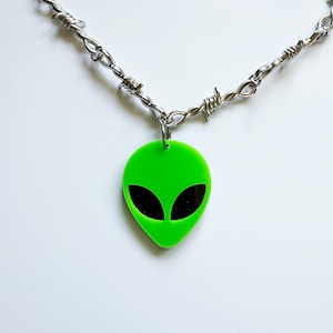 May include: A necklace featuring a bright neon green alien head pendant with black oval eyes. The pendant hangs from a silver-toned metal chain designed to resemble barbed wire, connected by a small circular jump ring at the top of the alien head.