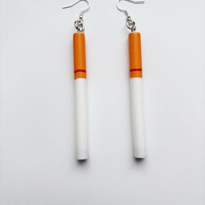 Op de afbeelding: Een paar novelty oorbellen ontworpen om op sigaretten te lijken. Elke oorbel heeft een witte cilindrische behuizing met een oranje punt en een rode band, opgehangen aan een zilverkleurige oorhaak.