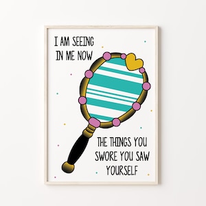 May include: A white background with a wooden frame around a print of a hand mirror with a turquoise and white striped surface. The mirror has a gold frame and pink beads around the edge. The mirror has a black handle and a gold heart at the top. The text "I am seeing in me now the things you swore you saw yourself" is printed below the mirror.