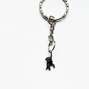 Puede incluir: Un llavero plateado con un pequeño dije de mono colgando de una cadena. El llavero tiene un anillo texturizado para sujetar las llaves. El dije de mono es detallado y parece estar hecho de metal.